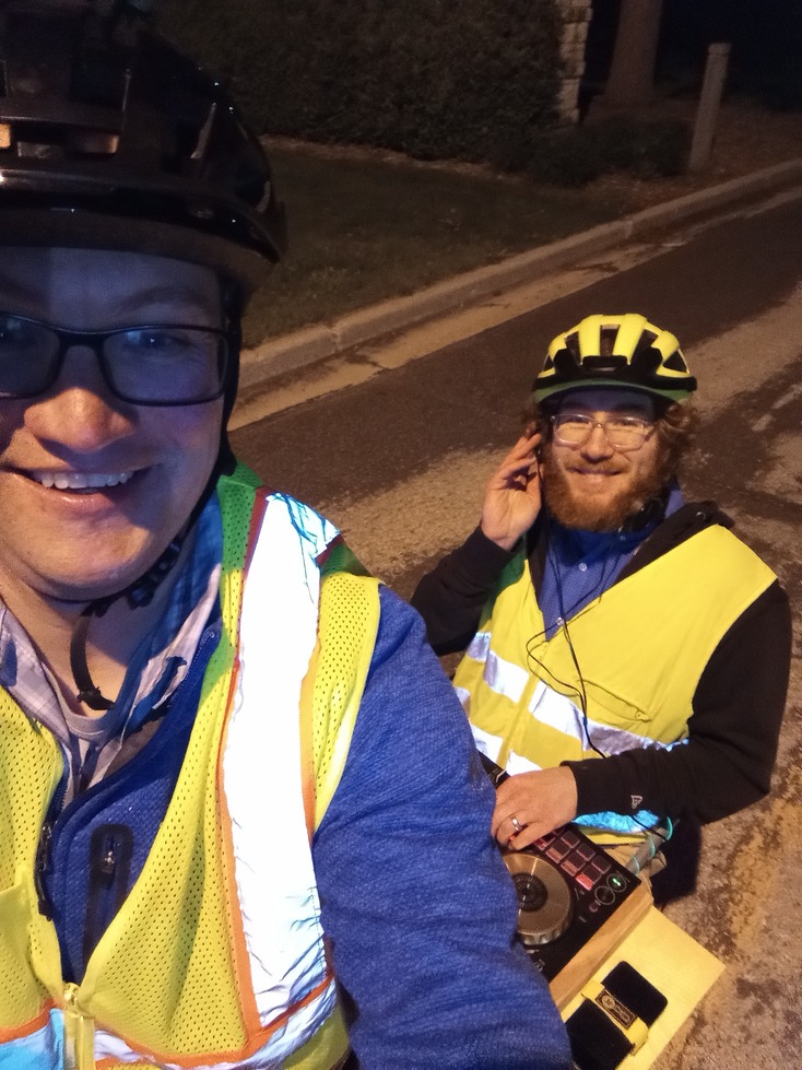 selfy of two men on a cargo bike, wearing reflective vests. The passenger is operating a dj controller